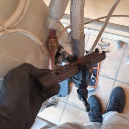 Strefa AC Sławomir Smoter - Instalator w czarnych rękawicach łączy rury klimatyzacji za pomocą specjalistycznego narzędzia, widoczne izolowane rury i fragment klimatyzatora.