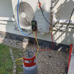 Strefa AC Sławomir Smoter - Zewnętrzna jednostka klimatyzacji Rotenso Hiro Series podłączona do manometru i butli z gazem chłodniczym podczas serwisu, widoczne przewody w kolorach czerwonym, żółtym i niebieskim. Na ścianie...