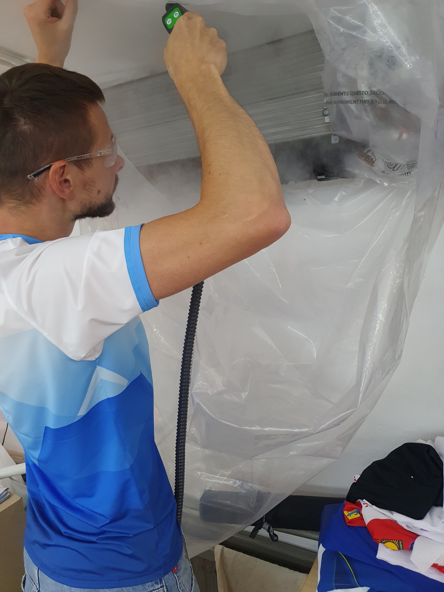 Mężczyzna w okularach ochronnych czyści klimatyzator zawieszony pod sufitem, używając specjalnego urządzenia z dyszą, całość zabezpieczona folią ochronną.
