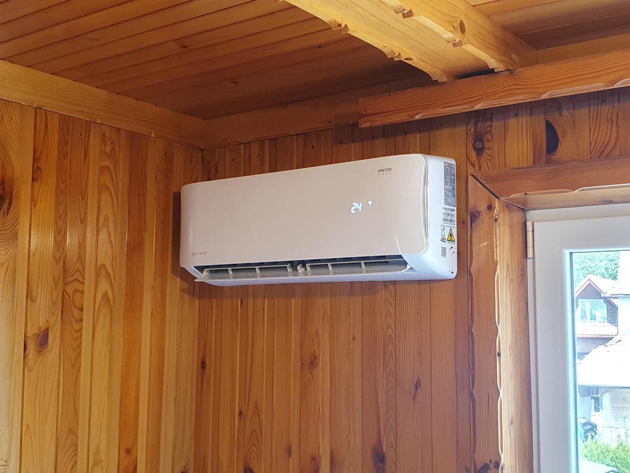 Biała jednostka wewnętrzna klimatyzatora marki Imoto zamontowana na drewnianej ścianie w domu z drewnianym sufitem, wyświetlająca temperaturę 24 stopnie Celsjusza.