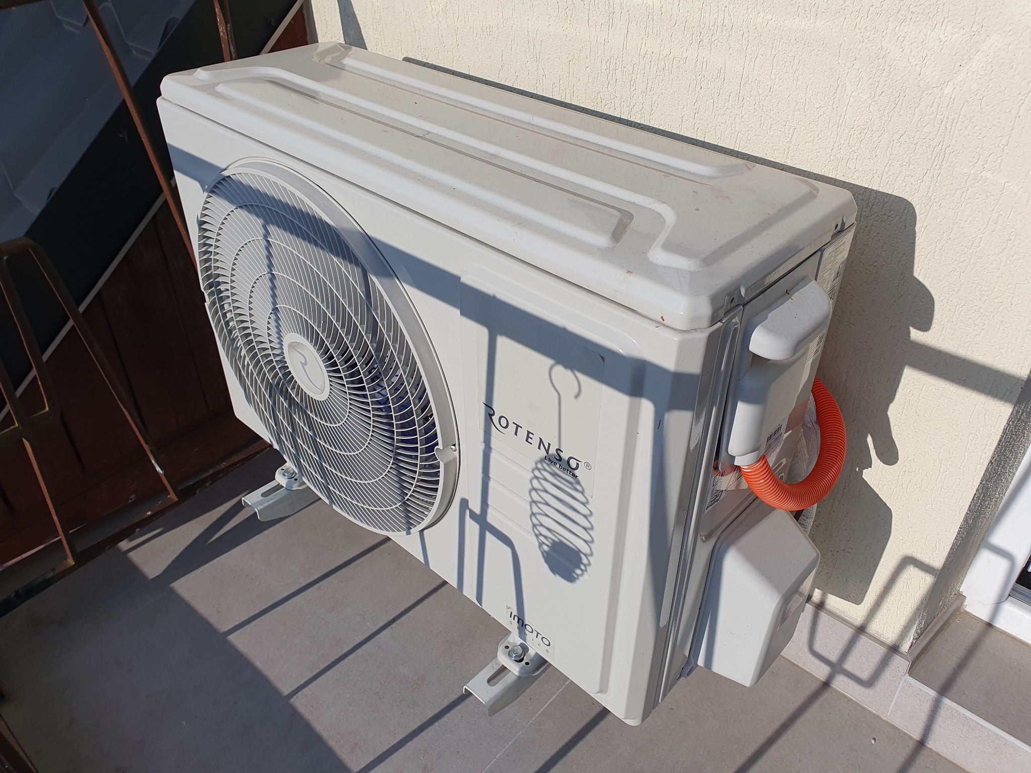 Zewnętrzna jednostka klimatyzacji Rotenso Imoto zamontowana na balkonie, widoczna pomarańczowa rurka odprowadzająca skropliny, cień rzucany przez kratkę balkonu.