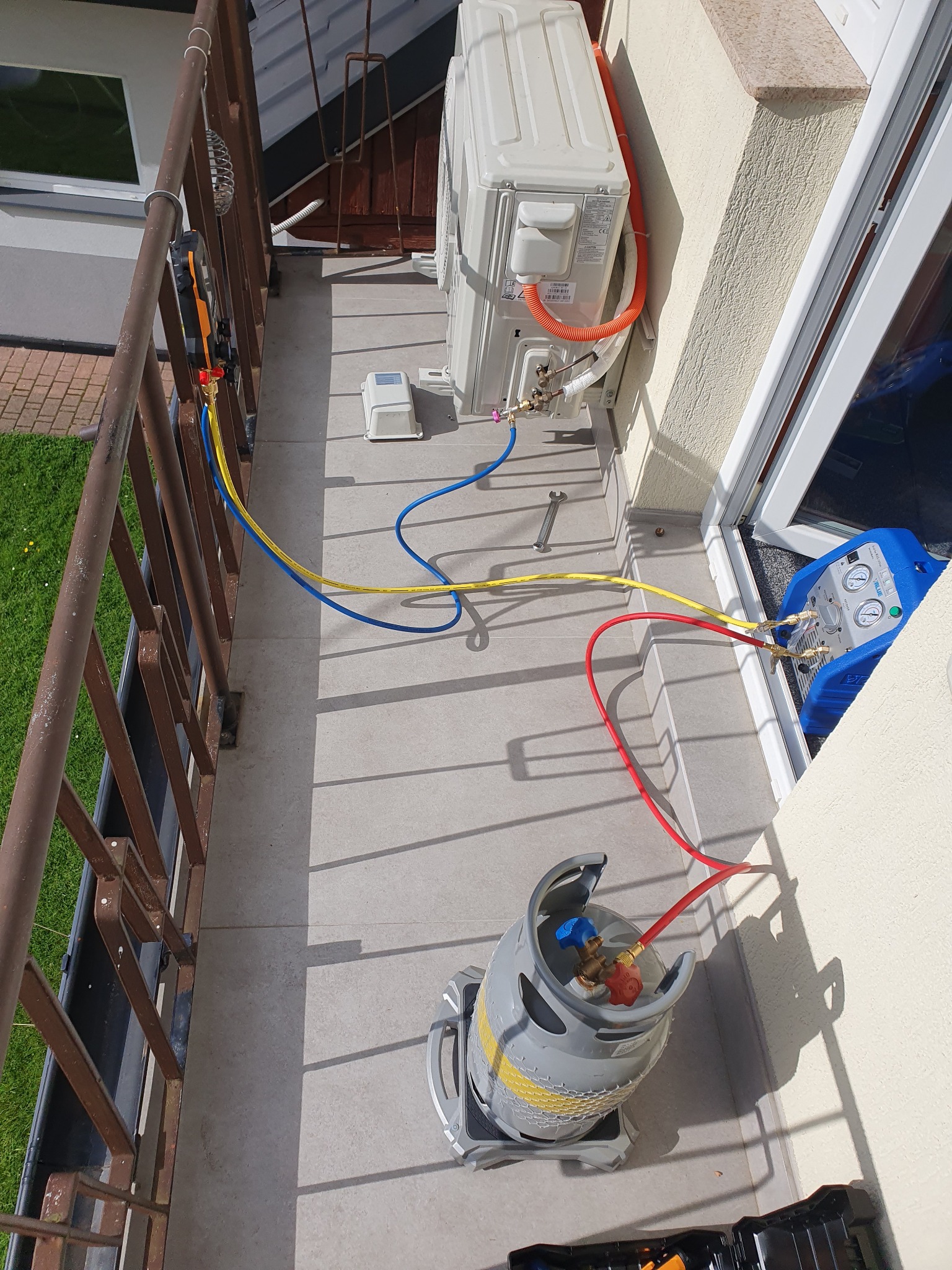 Montaż zewnętrznej jednostki klimatyzacji na balkonie, widoczne przewody, narzędzia i butla z gazem chłodniczym podczas podłączenia.