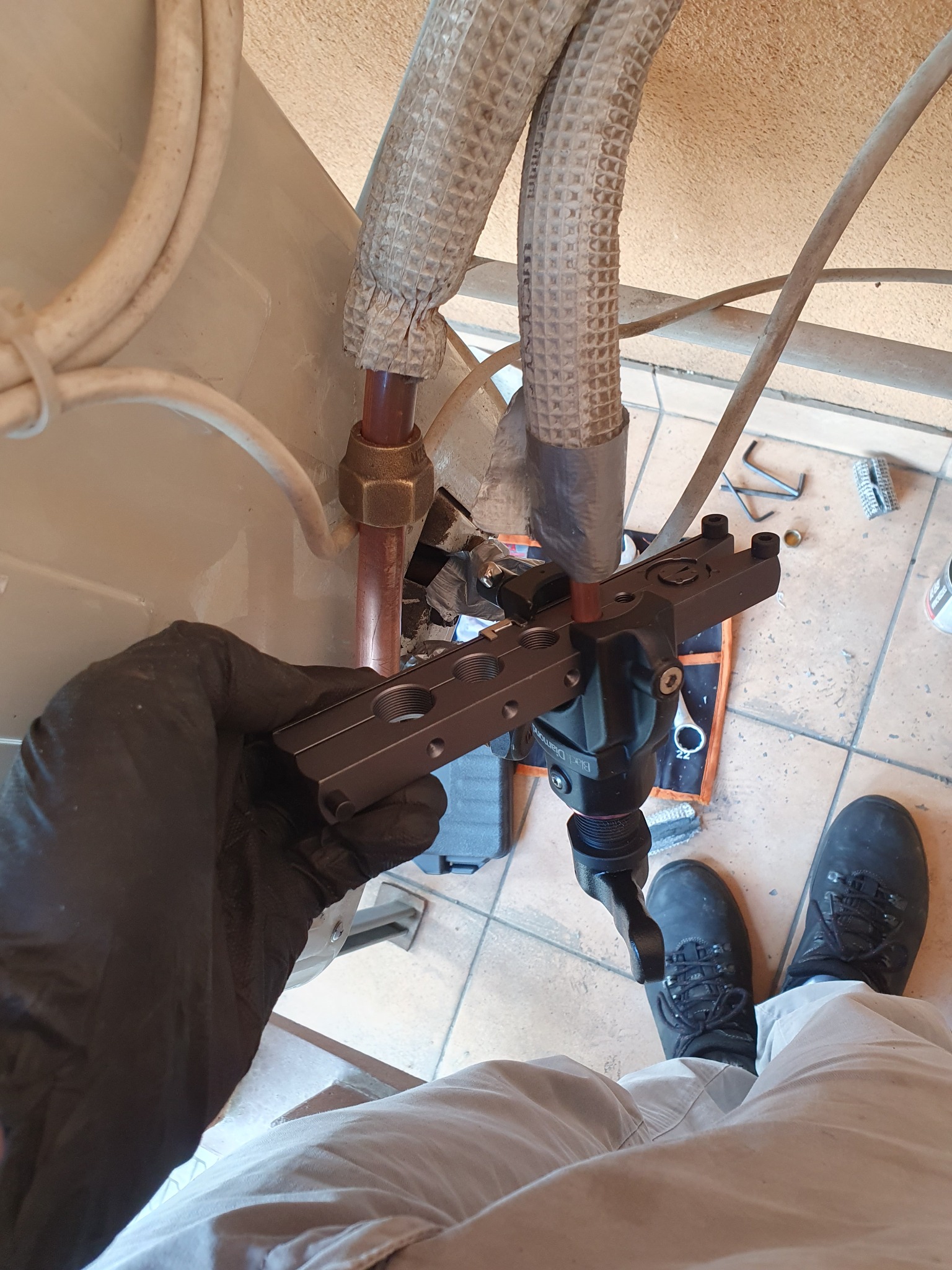 Instalator w czarnych rękawicach łączy rury klimatyzacji za pomocą specjalistycznego narzędzia, widoczne izolowane rury i fragment klimatyzatora.