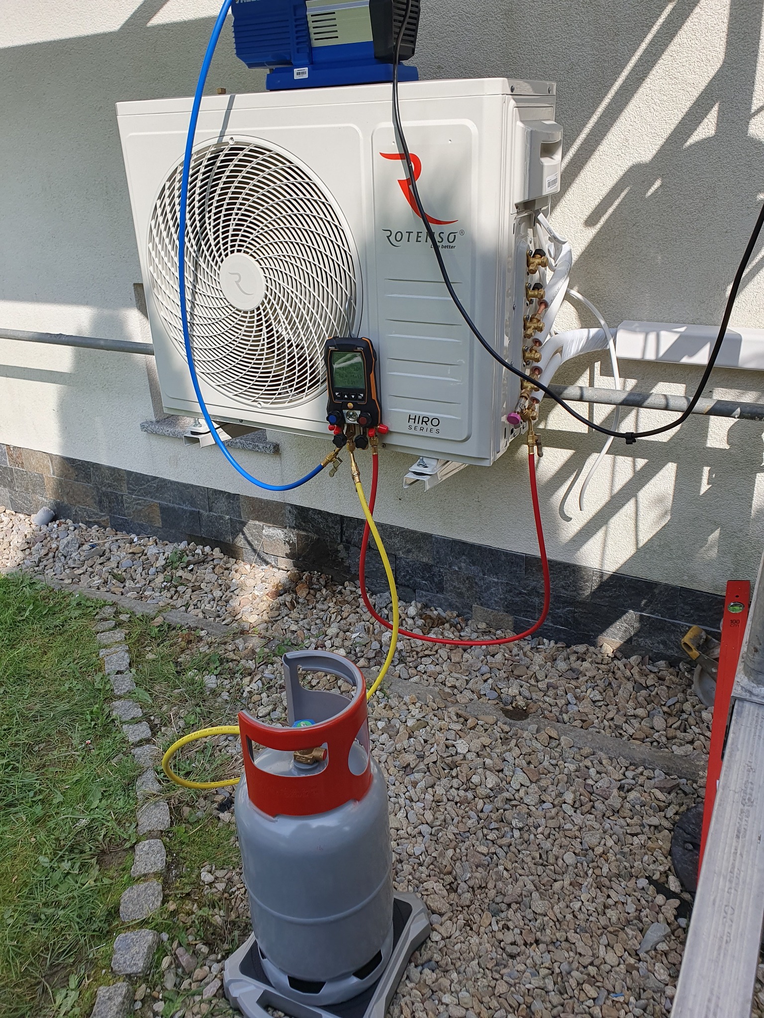 Zewnętrzna jednostka klimatyzacji Rotenso Hiro Series podłączona do manometru i butli z gazem chłodniczym podczas serwisu, widoczne przewody w kolorach czerwonym, żółtym i niebieskim. Na ścianie...