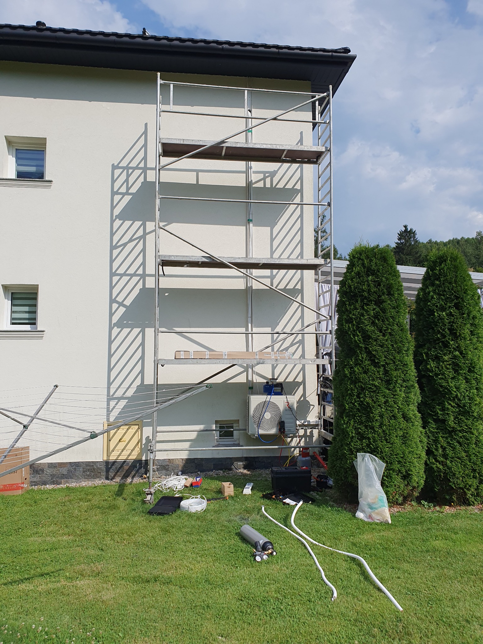 Montaż klimatyzacji na elewacji domu jednorodzinnego, widoczne rusztowanie, jednostka zewnętrzna klimatyzatora, narzędzia na trawniku i dwie wysokie tuje w tle.