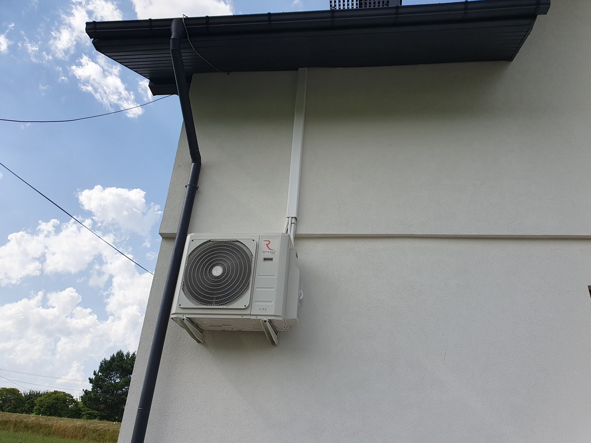 Zewnętrzna jednostka klimatyzacji Rotenso Hiro zamontowana na ścianie budynku z widoczną rurą odprowadzającą i czarną rynną dachową na tle błękitnego nieba z chmurami.