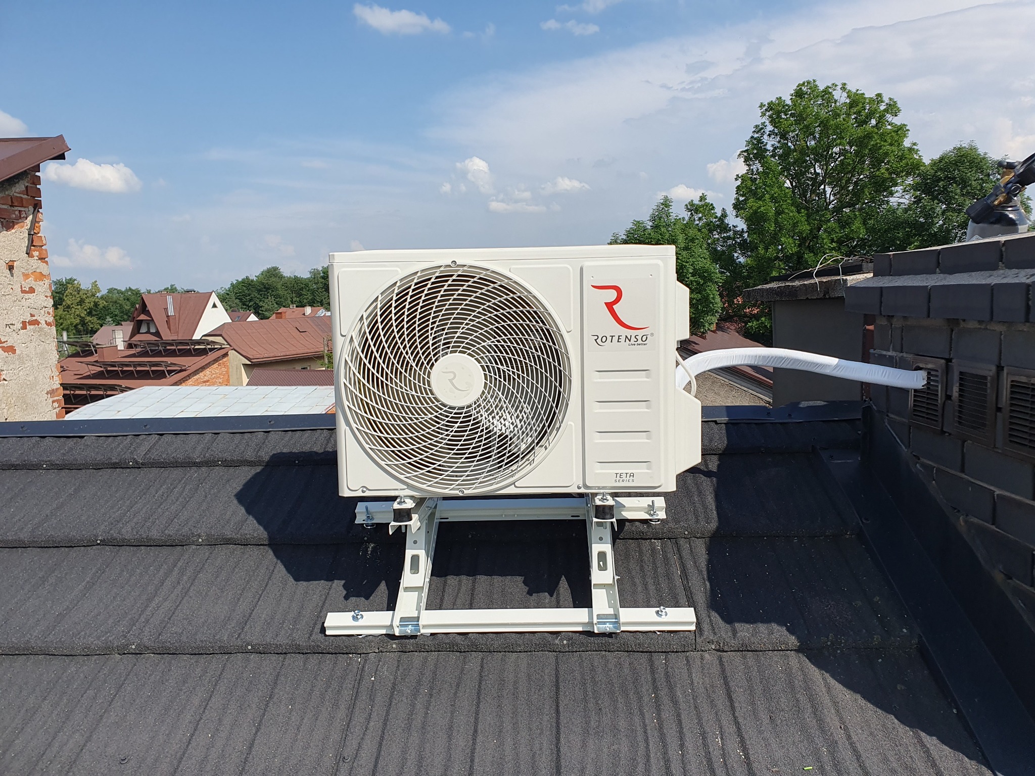 Zewnętrzna jednostka klimatyzacji Rotenso TETA Series zamontowana na dachu z czarnej dachówki, widok na okoliczne budynki i błękitne niebo z chmurami.