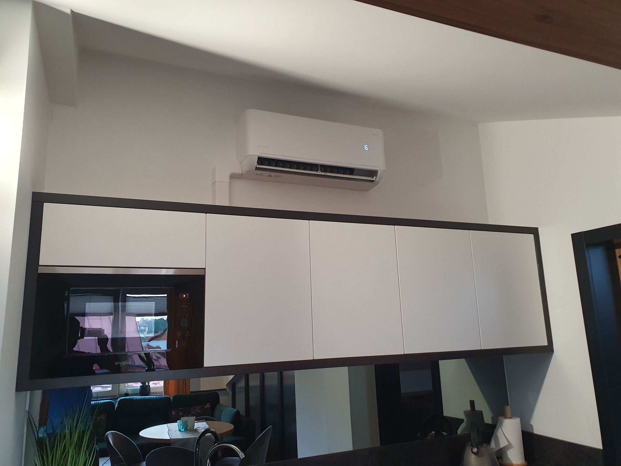 Biała jednostka klimatyzacyjna zamontowana nad rzędem białych szafek kuchennych z czarnymi ramami, widoczny fragment salonu w odbiciu.