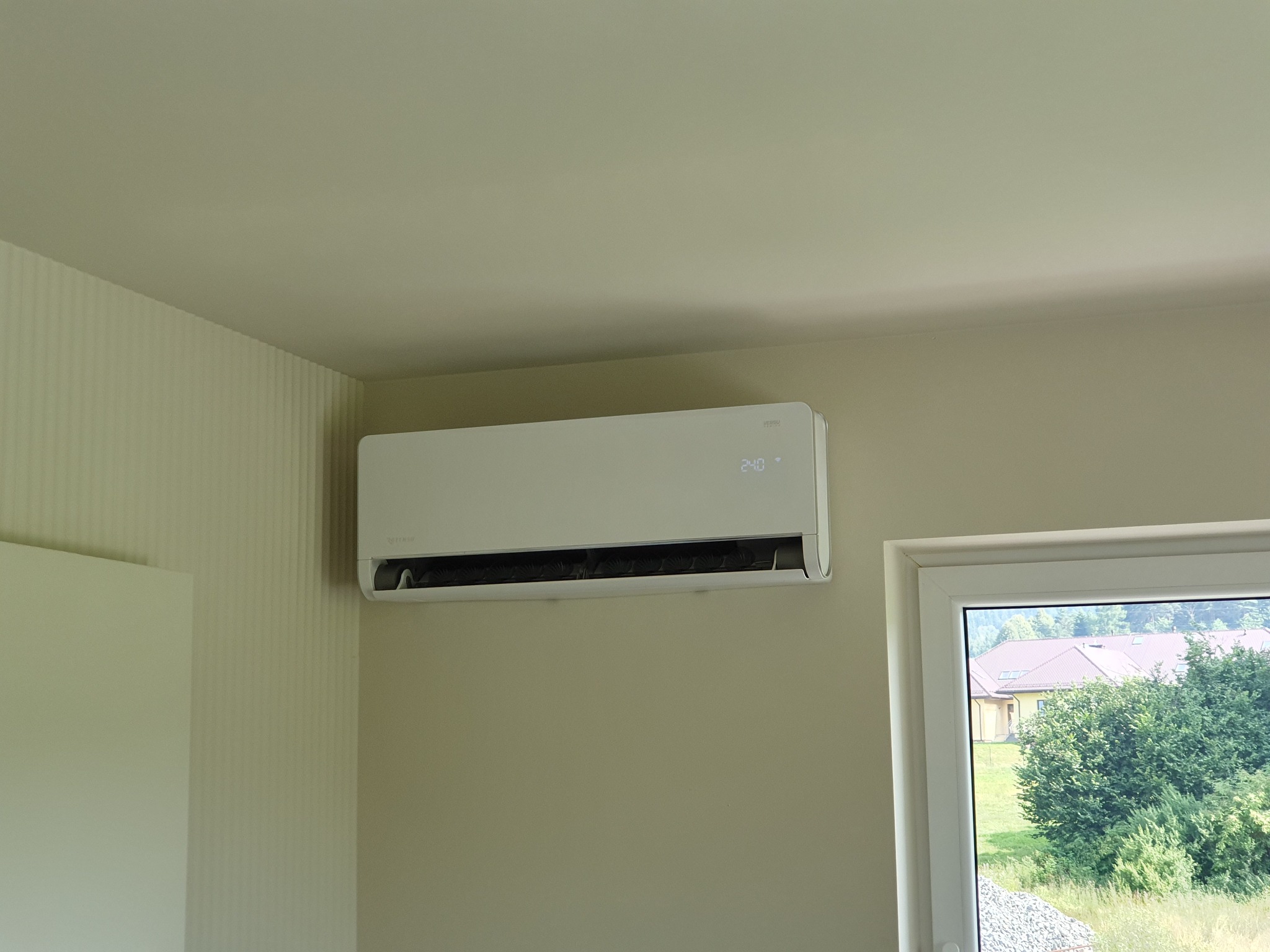 Biały klimatyzator ścienny zamontowany w jasnym pokoju, widok z okna na zieleń i zabudowania.