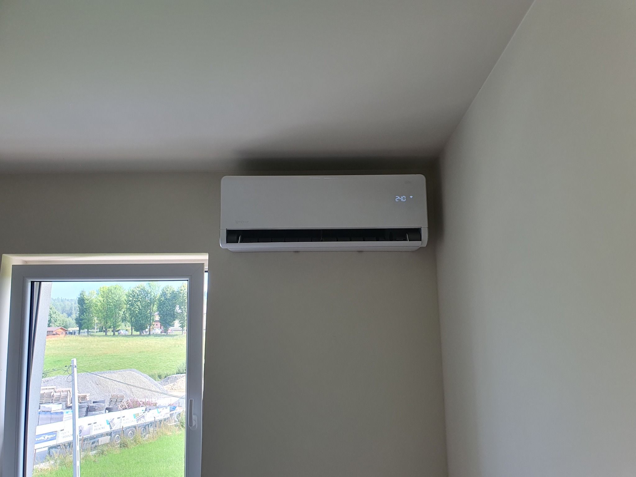 Biała jednostka klimatyzacji ściennej w jasnym pokoju, widok z okna na zielony krajobraz i plac budowy.