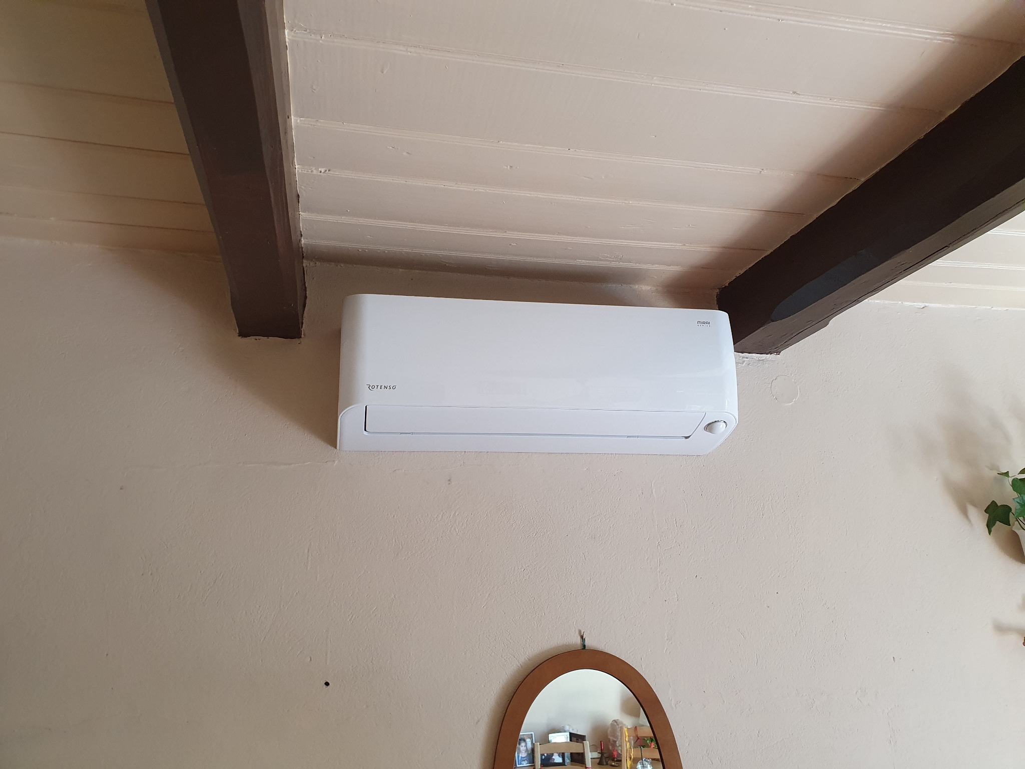 Biały klimatyzator marki Rotenso zamontowany na beżowej ścianie w pomieszczeniu z drewnianymi belkami stropowymi i dekoracyjnym lustrem.