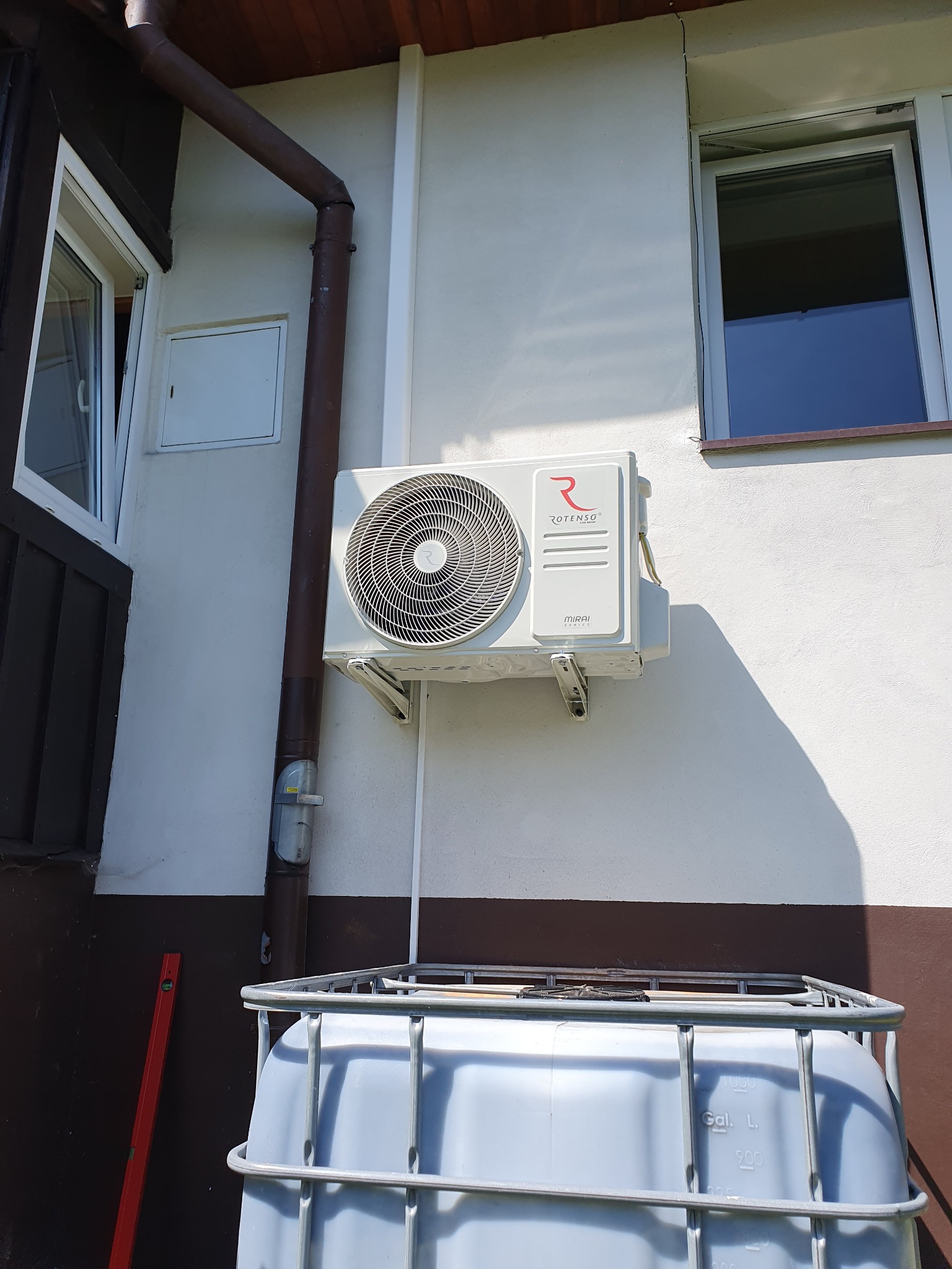 Zewnętrzna jednostka klimatyzacji Rotenso Mirai zamontowana na białej elewacji domu, widoczna rynna i okno, na pierwszym planie zbiornik na wodę w metalowej klatce.