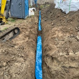 Instalacje Elektryczne - Wykop pod fundamenty zabezpieczony niebieską folią, widoczny mini-koparka, toaleta przenośna i pracownik z łopatą w tle.