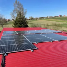 Panele fotowoltaiczne zamontowane na czerwonym, blachodachówkowym dachu, widok z góry na tle zielonych pól i błękitnego nieba.