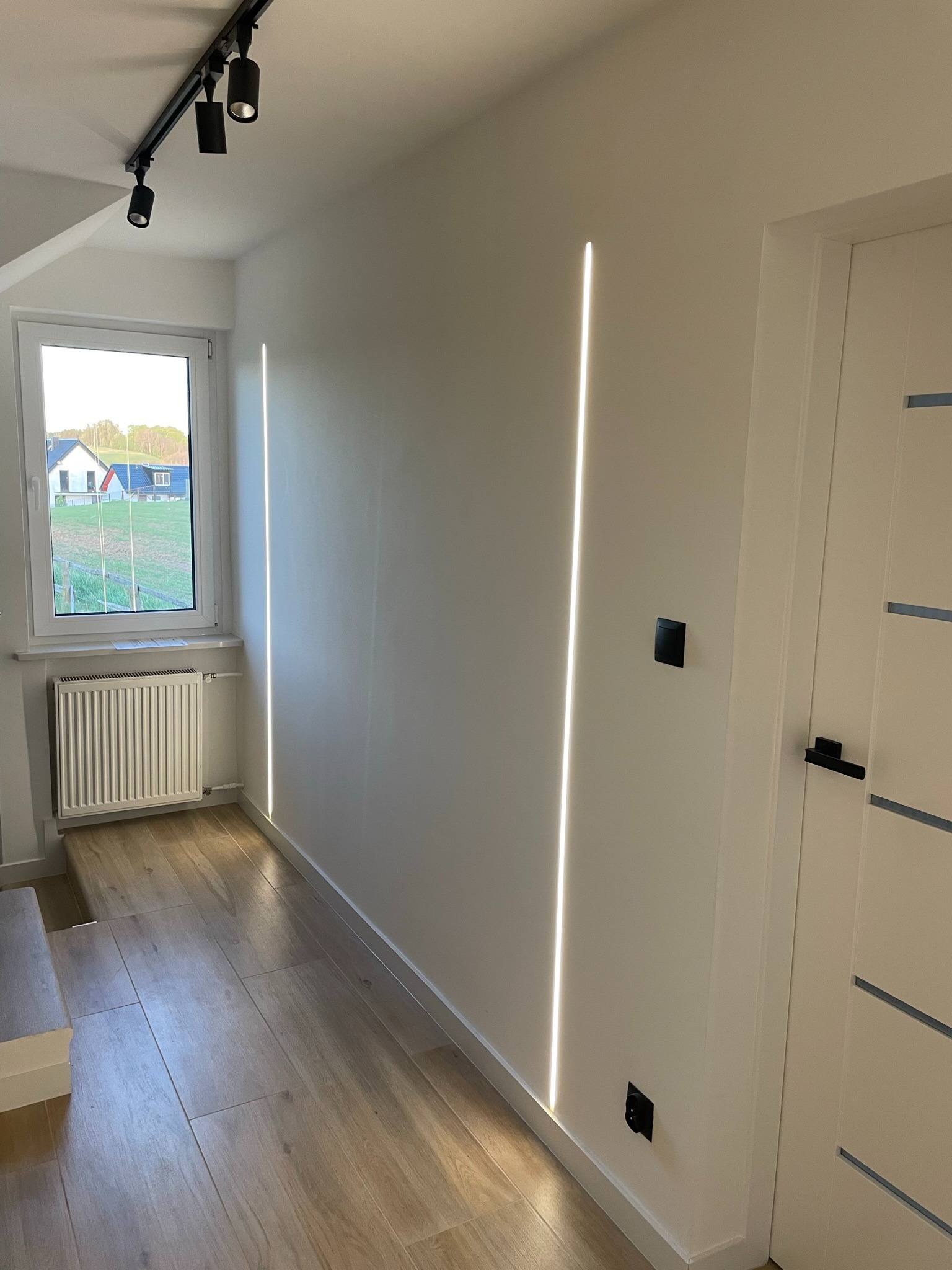 Nowoczesne oświetlenie korytarza z dwiema pionowymi liniami światła w ścianie, uzupełnione reflektorami na szynie sufitowej, widok na zewnątrz przez okno.