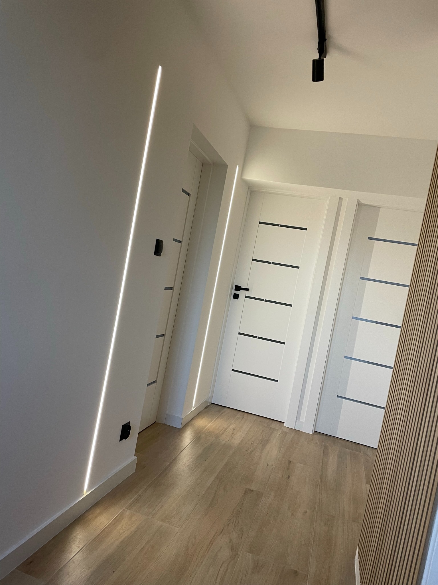 Nowoczesny korytarz z podświetleniem LED wzdłuż ściany, jasnymi drzwiami z poziomymi wstawkami i drewnianą okładziną ścienną.