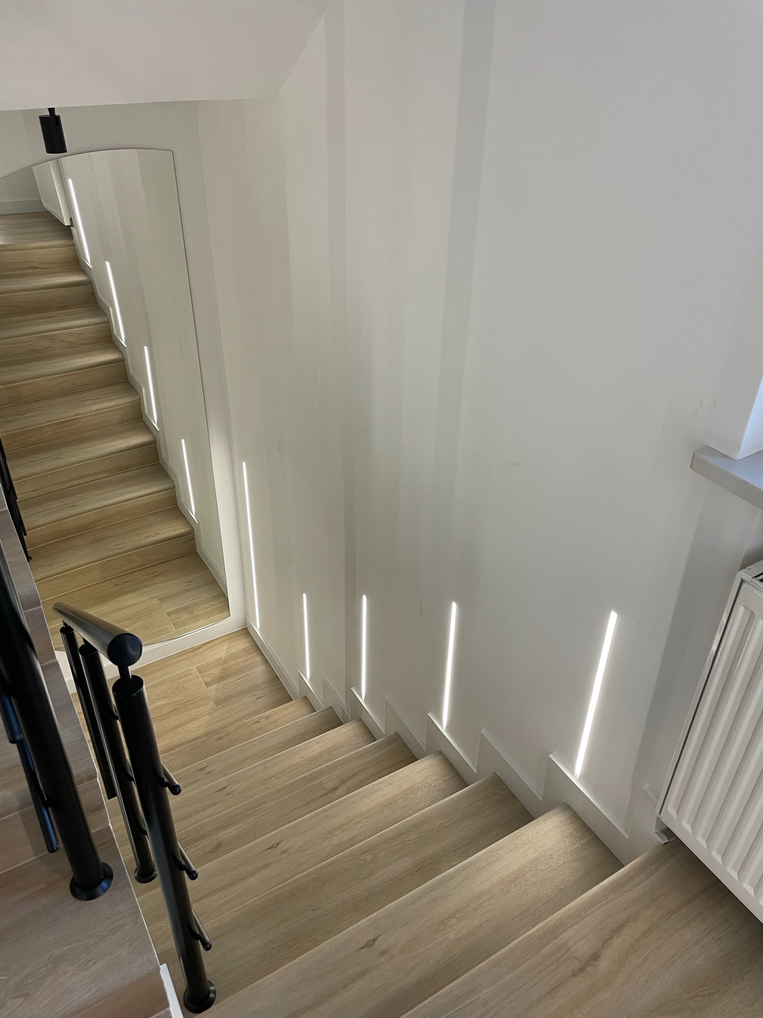 Nowoczesne schody z drewnopodobnymi stopniami, oświetleniem LED w ścianie i czarną metalową balustradą, widok z góry.