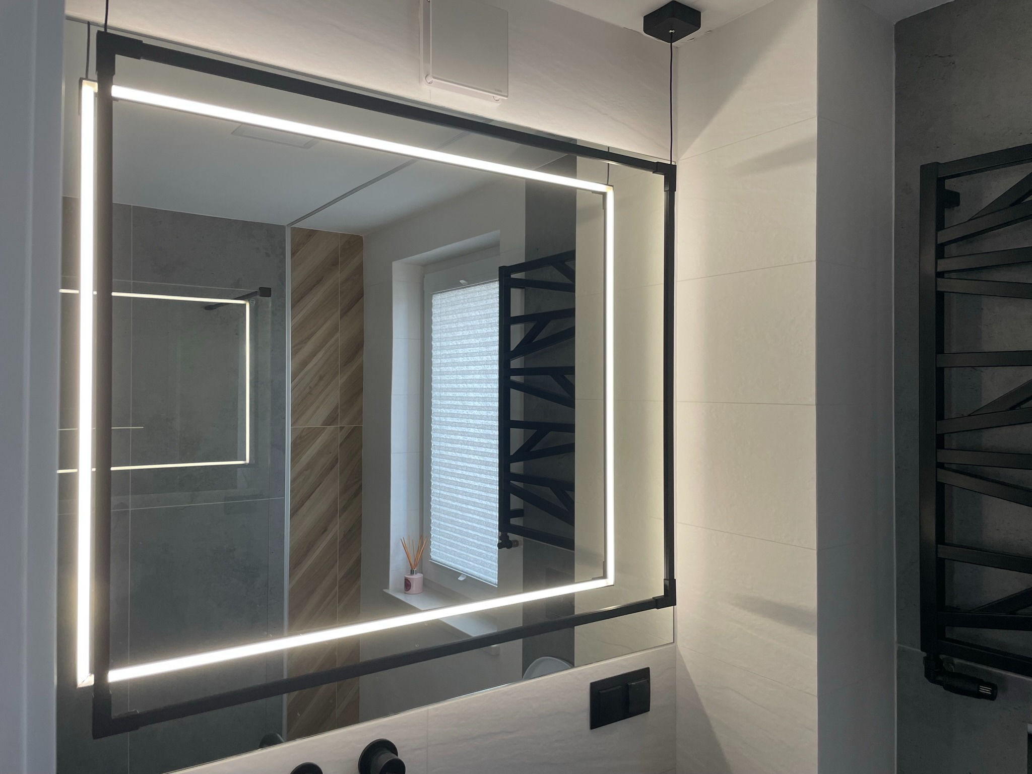 Nowoczesna łazienka z podświetlanym lustrem w czarnej ramie, odbijającym fragment prysznica i okno z roletami, czarny grzejnik drabinkowy na ścianie.