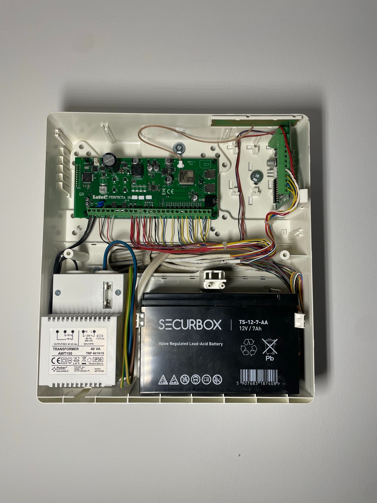 Otwarta obudowa centrali alarmowej z widoczną elektroniką, baterią akumulatorową i transformatorem, liczne przewody połączeniowe.