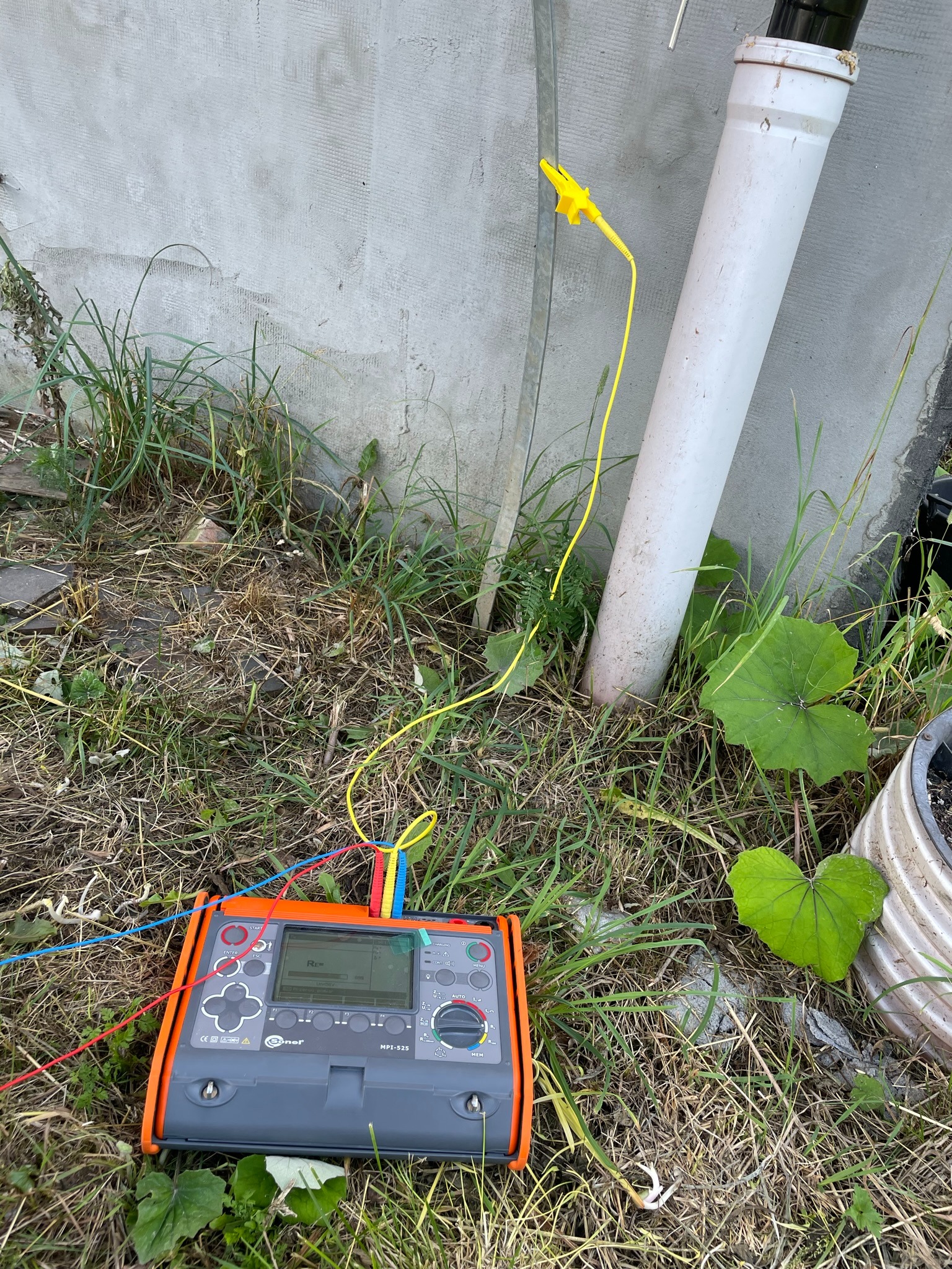 Miernik elektryczny MPI-525 z podłączonymi przewodami pomiarowymi w trawie, obok betonowej ściany i rury PCV, żółty zacisk na metalowej rurze.