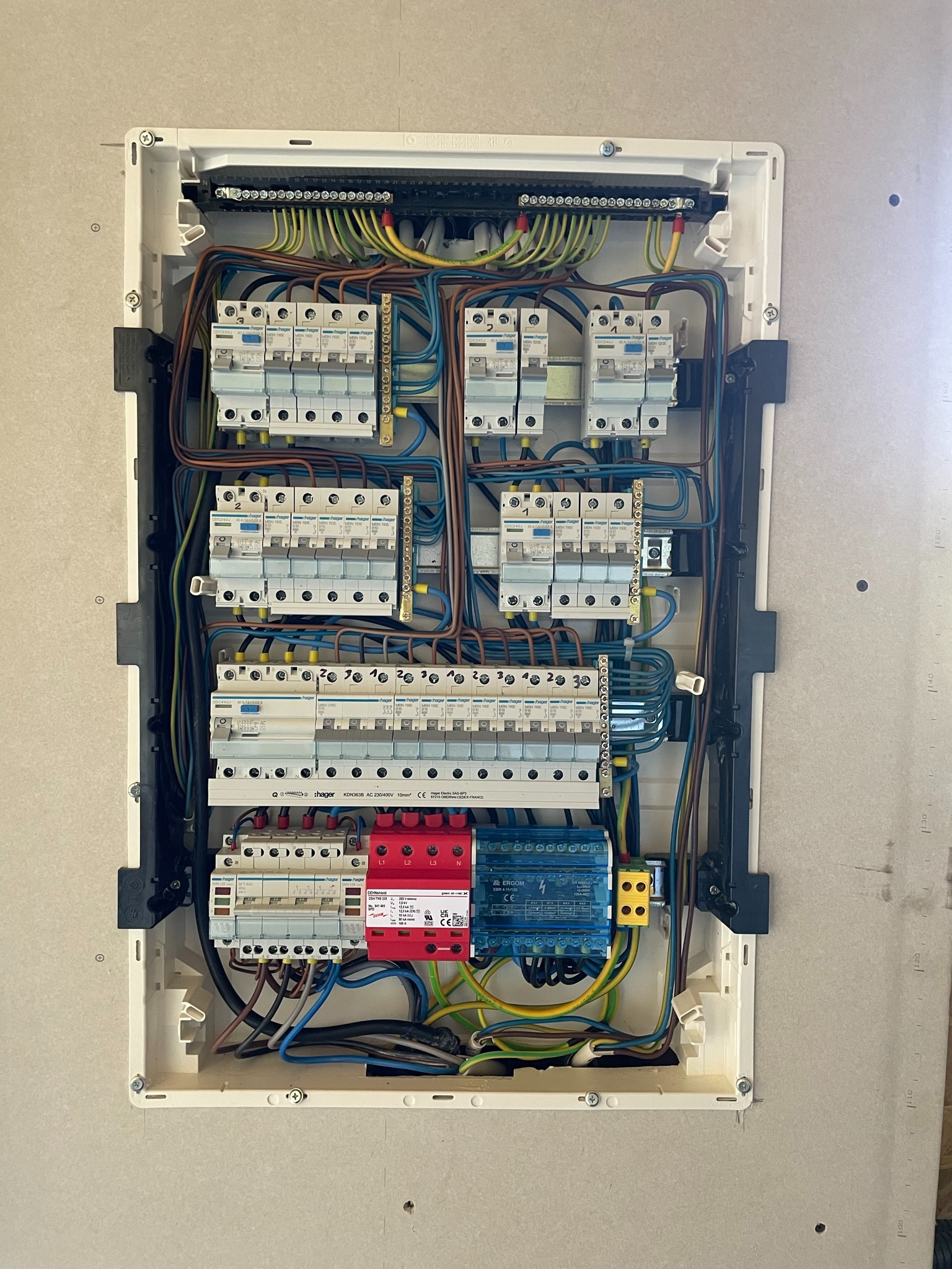Rozdzielnica elektryczna w obudowie, widoczne zabezpieczenia nadprądowe, bezpieczniki, przewody w różnych kolorach, listwy zaciskowe. Elementy marki Hager i Dehn. Widok z góry na zamontowaną...
