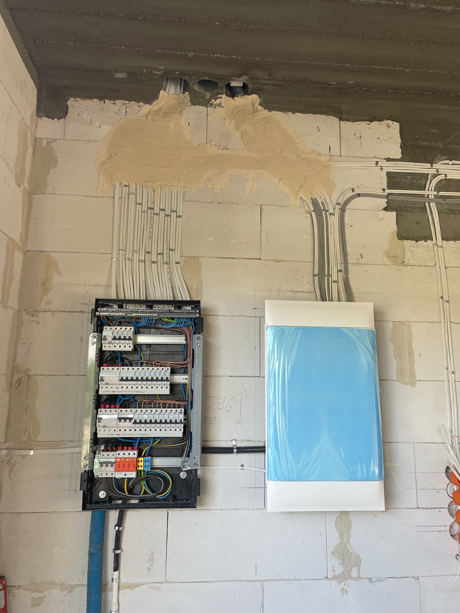 Rozdzielnica elektryczna z podłączonymi przewodami na surowej ścianie z betonu komórkowego, obok nowy grzejnik w folii ochronnej, widoczne ślady po tynku.