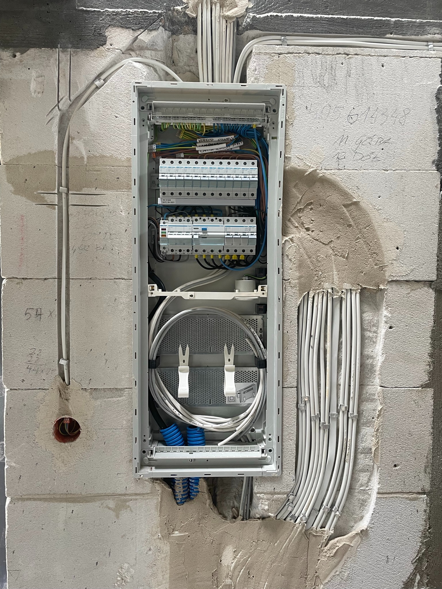 Rozdzielnica elektryczna w trakcie montażu w surowej ścianie z betonu komórkowego, widoczne kable w peszlach i listwach, zabezpieczenia różnicowoprądowe i nadprądowe oraz przewody światłowodowe.