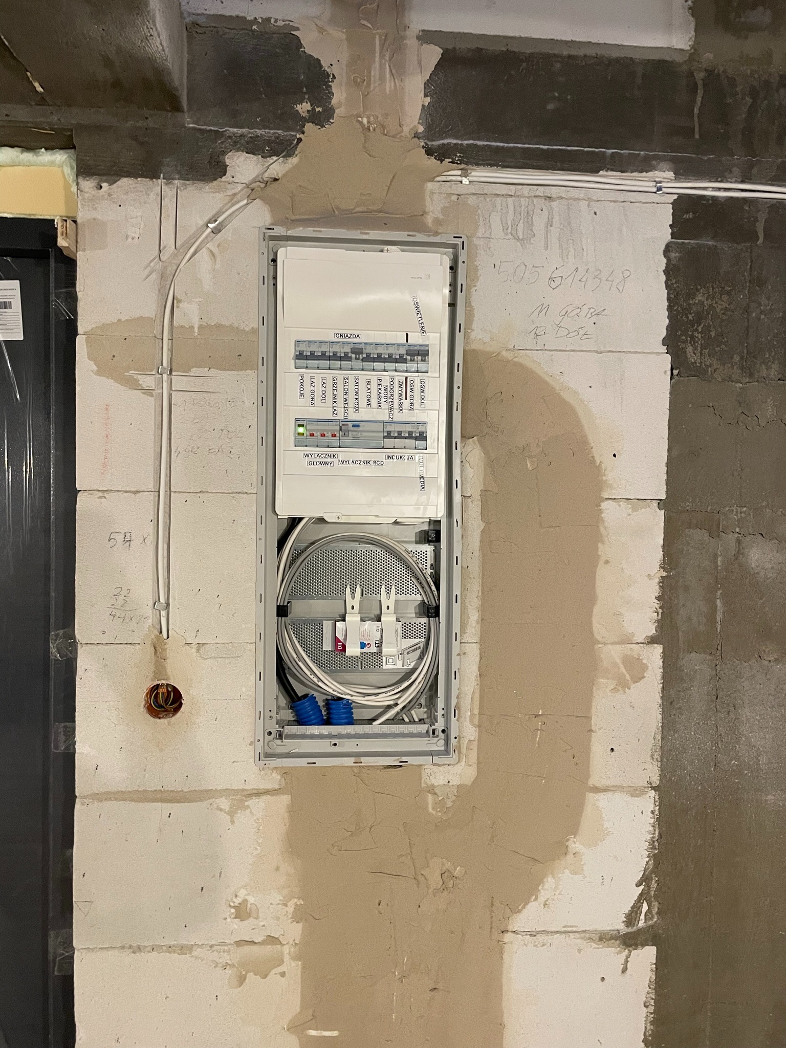 Szafka elektryczna z okablowaniem w trakcie montażu, widoczna na surowej ścianie w Staniszewie, z otworami na kable i widocznymi przewodami elektrycznymi.