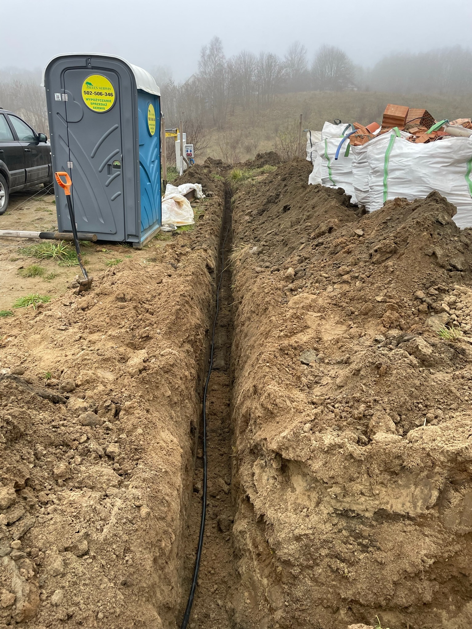 Wykopany rów w ziemi z ułożonym kablem, obok przenośna toaleta i sterta cegieł na placu budowy w pochmurny dzień.