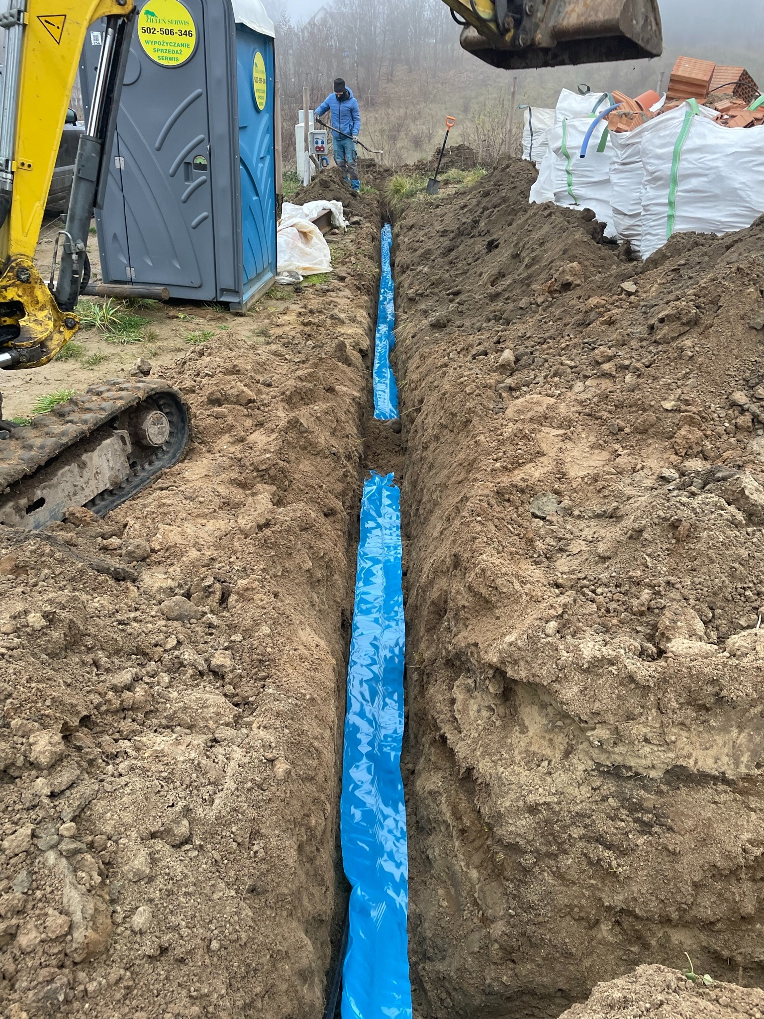 Wykop pod fundamenty zabezpieczony niebieską folią, widoczny mini-koparka, toaleta przenośna i pracownik z łopatą w tle.