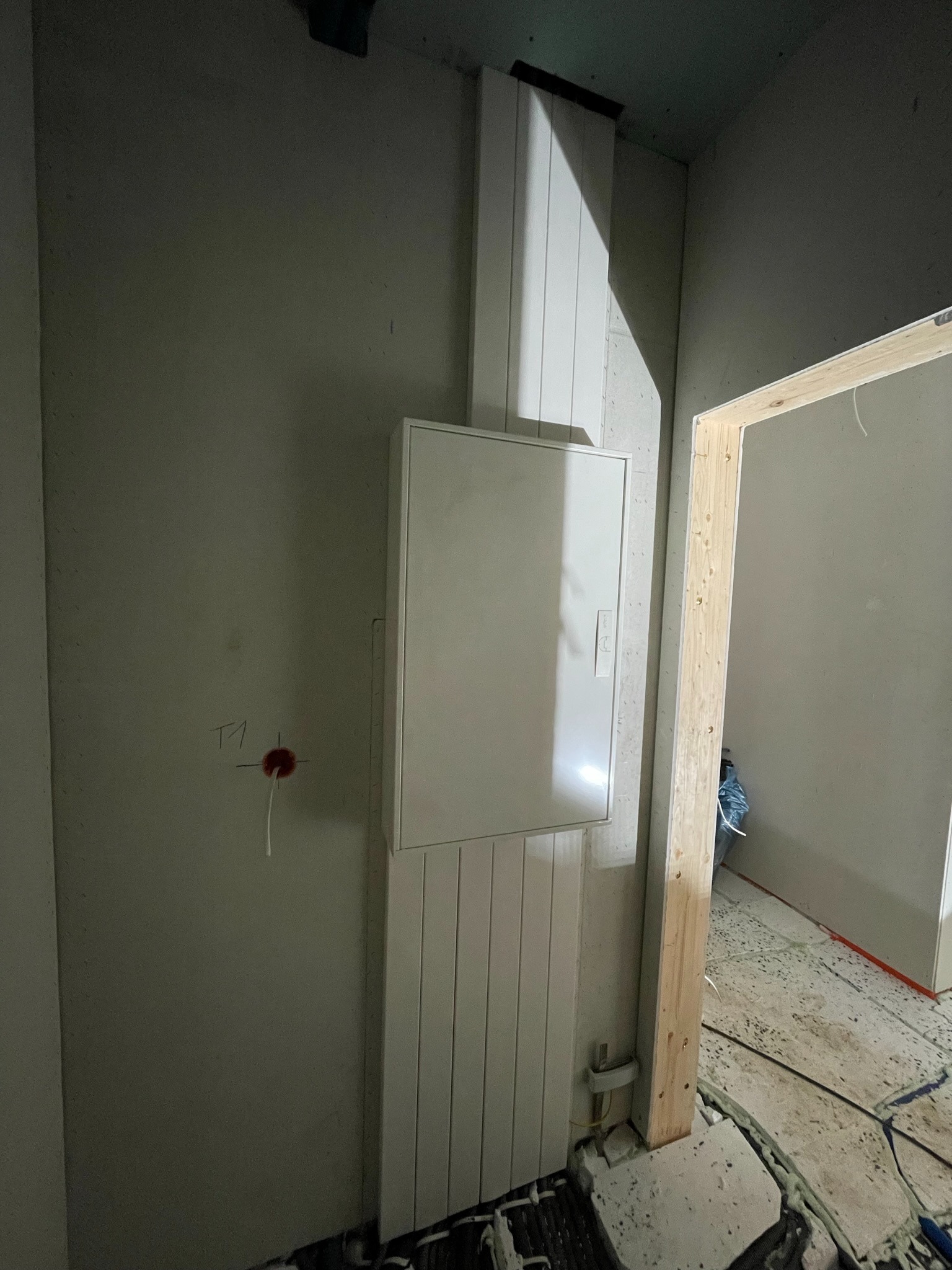 Biała skrzynka elektryczna zamontowana na ścianie w surowym wnętrzu, widoczne przewody i otwór na gniazdko, fragment ościeżnicy drzwiowej w tle.