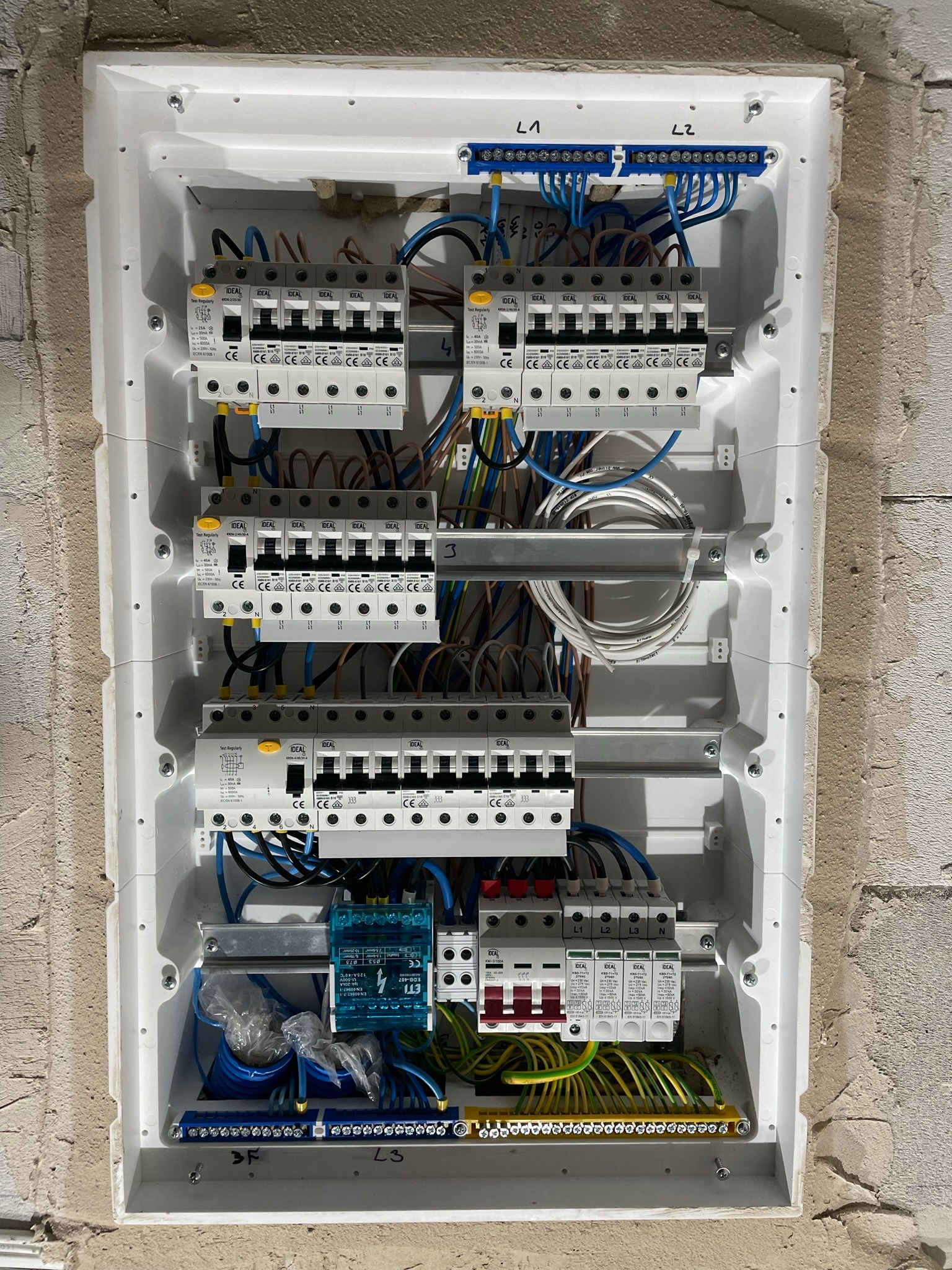 Rozdzielnica elektryczna z zamontowanymi bezpiecznikami i okablowaniem w białej obudowie, widok z góry, surowa ściana w tle.