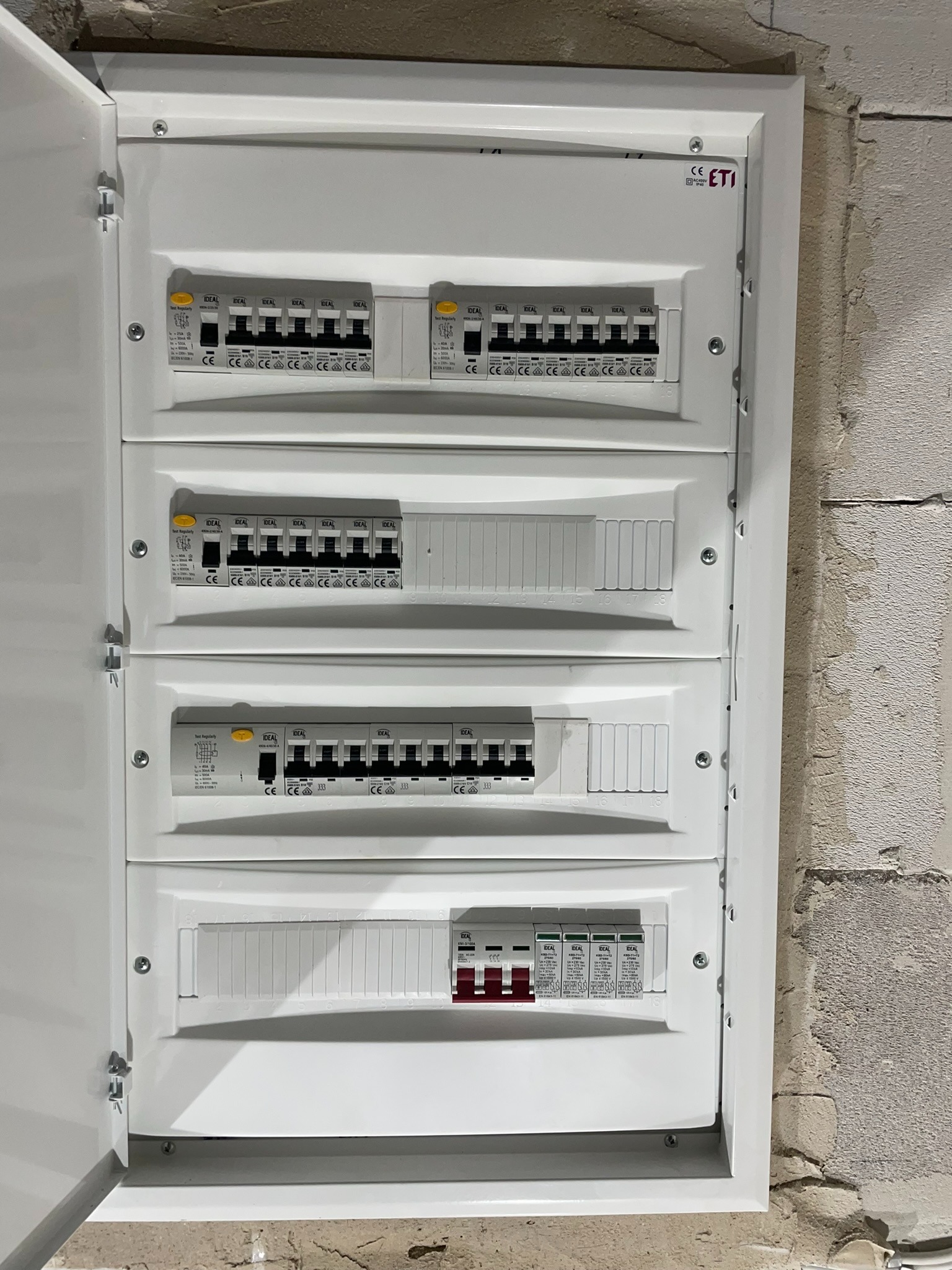Otwarta skrzynka elektryczna z czterema rzędami zabezpieczeń różnicowoprądowych i nadprądowych marki IDEAL, zamontowana na surowej ścianie z betonu komórkowego.