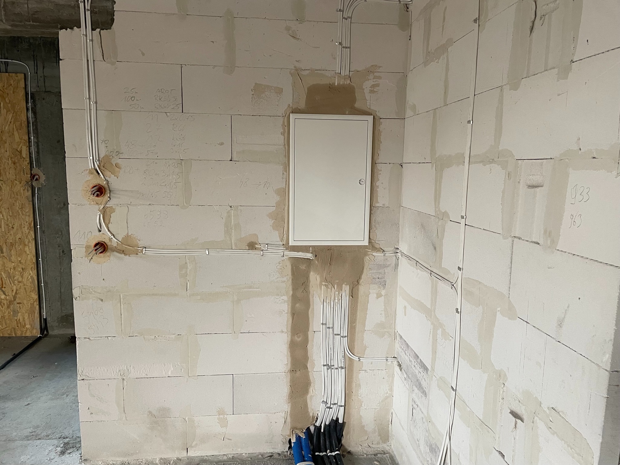 Białe przewody elektryczne prowadzone po ścianie z bloczków betonowych, z widoczną skrzynką rozdzielczą i otworami pod gniazdka, ściana z widocznymi notatkami technicznymi.