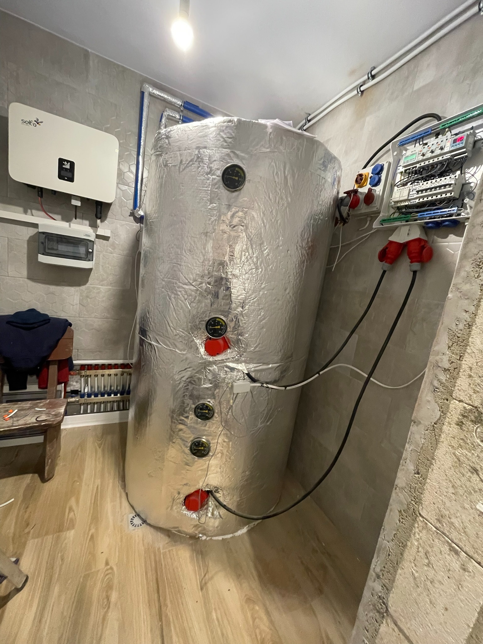 Instalacja zbiornika akumulacyjnego ciepła, owiniętego folią izolacyjną, z podłączonymi przewodami elektrycznymi i manometrami, widoczna skrzynka elektryczna z bezpiecznikami i falownik solarny...