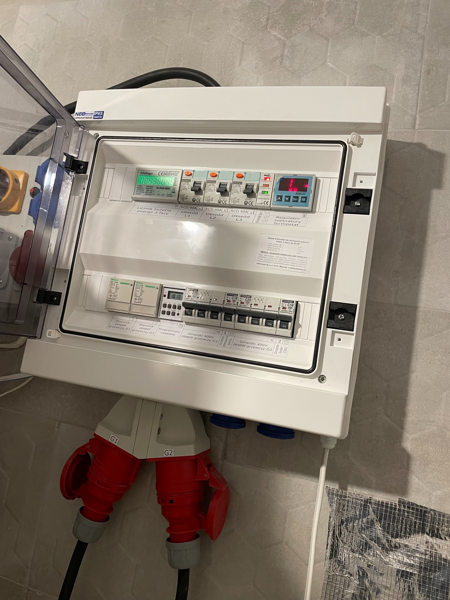 Rozdzielnica elektryczna z otwartą obudową, widoczne bezpieczniki, liczniki i regulatory, z podłączonymi czerwonymi gniazdami 400V, zamontowana na szarej ścianie z płytek.
