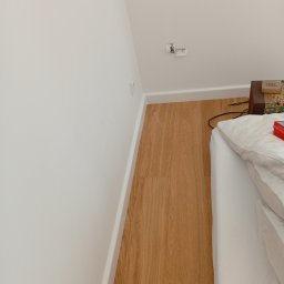 Usługi remontowo budowlane Sufity i wykończenia - Jasne panele podłogowe ułożone wzdłuż białej ściany z listwą przypodłogową. Fragment łóżka z białą pościelą w prawym dolnym rogu. Widok z góry, naturalne światło.