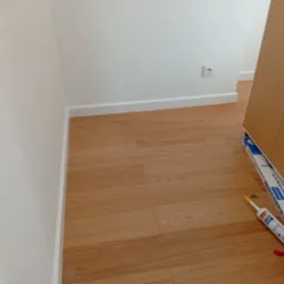 Jasne panele podłogowe po montażu, białe listwy przypodłogowe, silikon i uchwyt do TV. Widok z góry, ukończona praca.