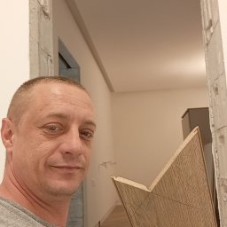 Usługi remontowo budowlane Sufity i wykończenia - Mężczyzna trzyma panel podłogowy z narysowaną linią cięcia na tle odnowionego wnętrza z widoczną podłogą. Fragment futryny w trakcie prac wykończeniowych.