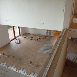 Usługi remontowo budowlane Sufity i wykończenia - Układanie parkietu w jodełkę w przestronnym, jasnym wnętrzu. Widoczne narzędzia, klej i częściowo ułożona podłoga. Ujęcie z góry, z widokiem na okno i schody.