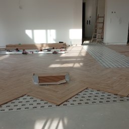 Usługi remontowo budowlane Sufity i wykończenia - Układanie parkietu w jodełkę. Widoczne panele, podkład i narzędzia. Częściowo ułożona podłoga w jasnym pomieszczeniu z dużym oknem i drabiną w tle.