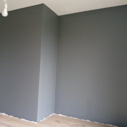 Usługi remontowo budowlane Sufity i wykończenia - Świeżo pomalowany pokój w odcieniach szarości, z widoczną żarówką i fragmentem drewnianej podłogi. Minimalistyczne wnętrze po remoncie.