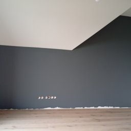 Usługi remontowo budowlane Sufity i wykończenia - Ściana pomalowana na szaro z widocznymi otworami na gniazdka elektryczne i fragmentem podłogi z paneli. Biały sufit z nietypowym załamaniem.