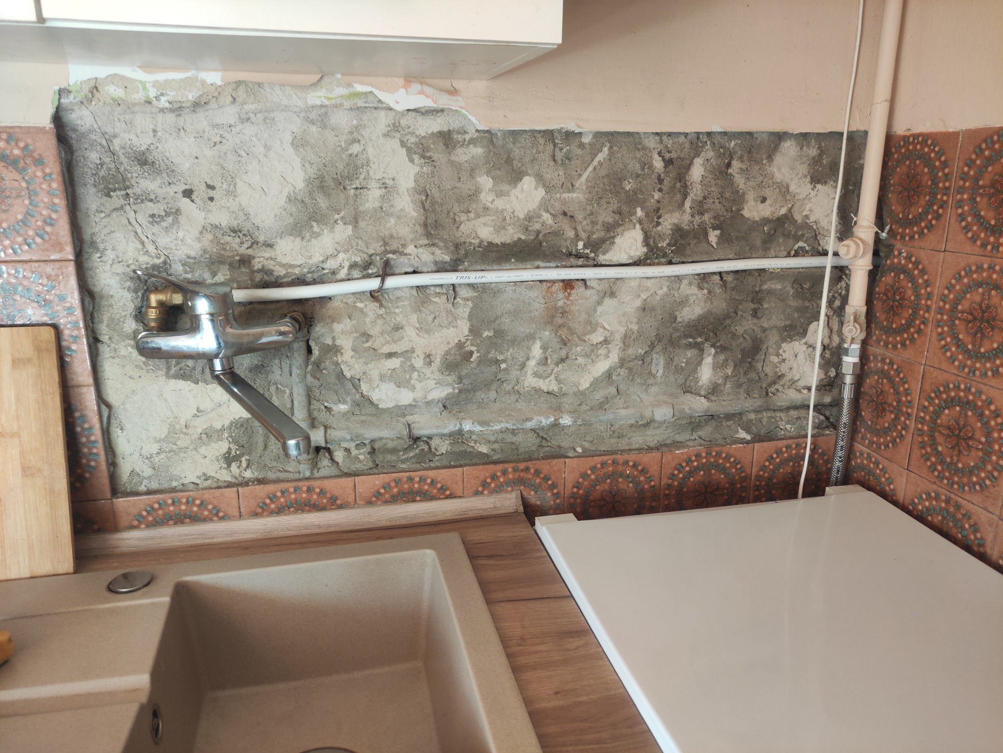 Niedokończona ściana w kuchni z odsłoniętym murem i fragmentami starych płytek, widoczna bateria kuchenna i rury instalacyjne.