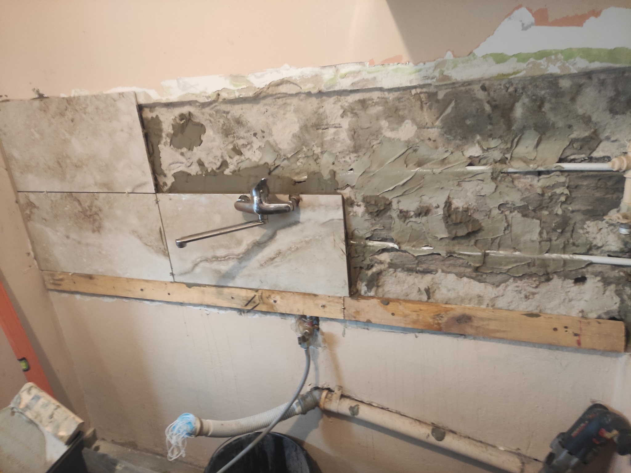 Fragment ściany w trakcie remontu z częściowo położonymi płytkami, widoczne rury i drewniane listwy podtrzymujące, pozostałości starej farby i zaprawy, oraz narzędzia w tle.