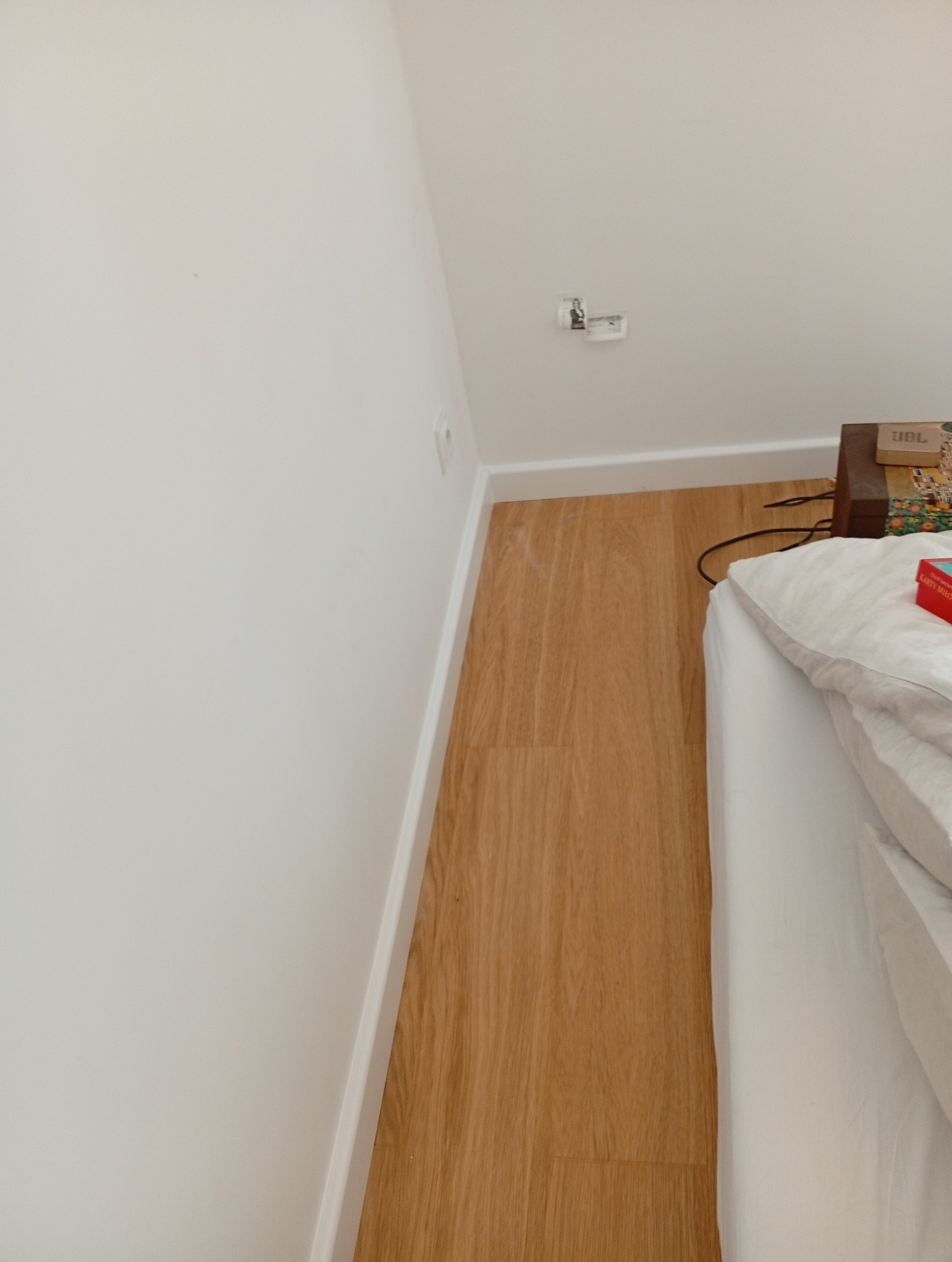 Jasne panele podłogowe ułożone wzdłuż białej ściany z listwą przypodłogową. Fragment łóżka z białą pościelą w prawym dolnym rogu. Widok z góry, naturalne światło.