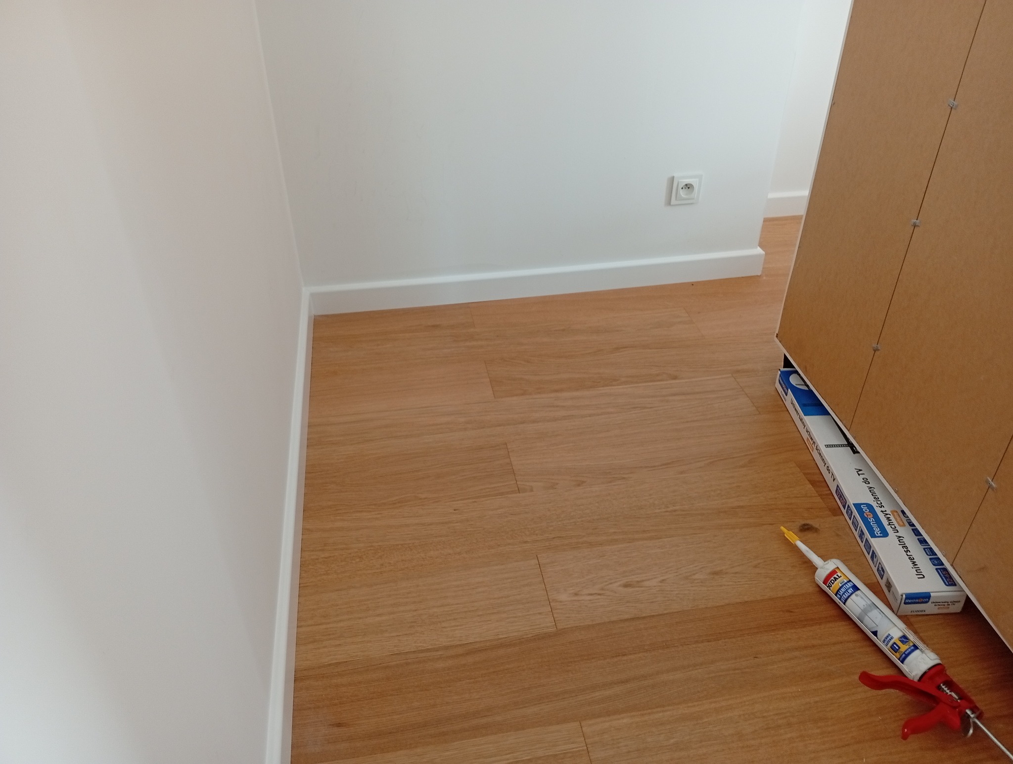 Jasne panele podłogowe po montażu, białe listwy przypodłogowe, silikon i uchwyt do TV. Widok z góry, ukończona praca.