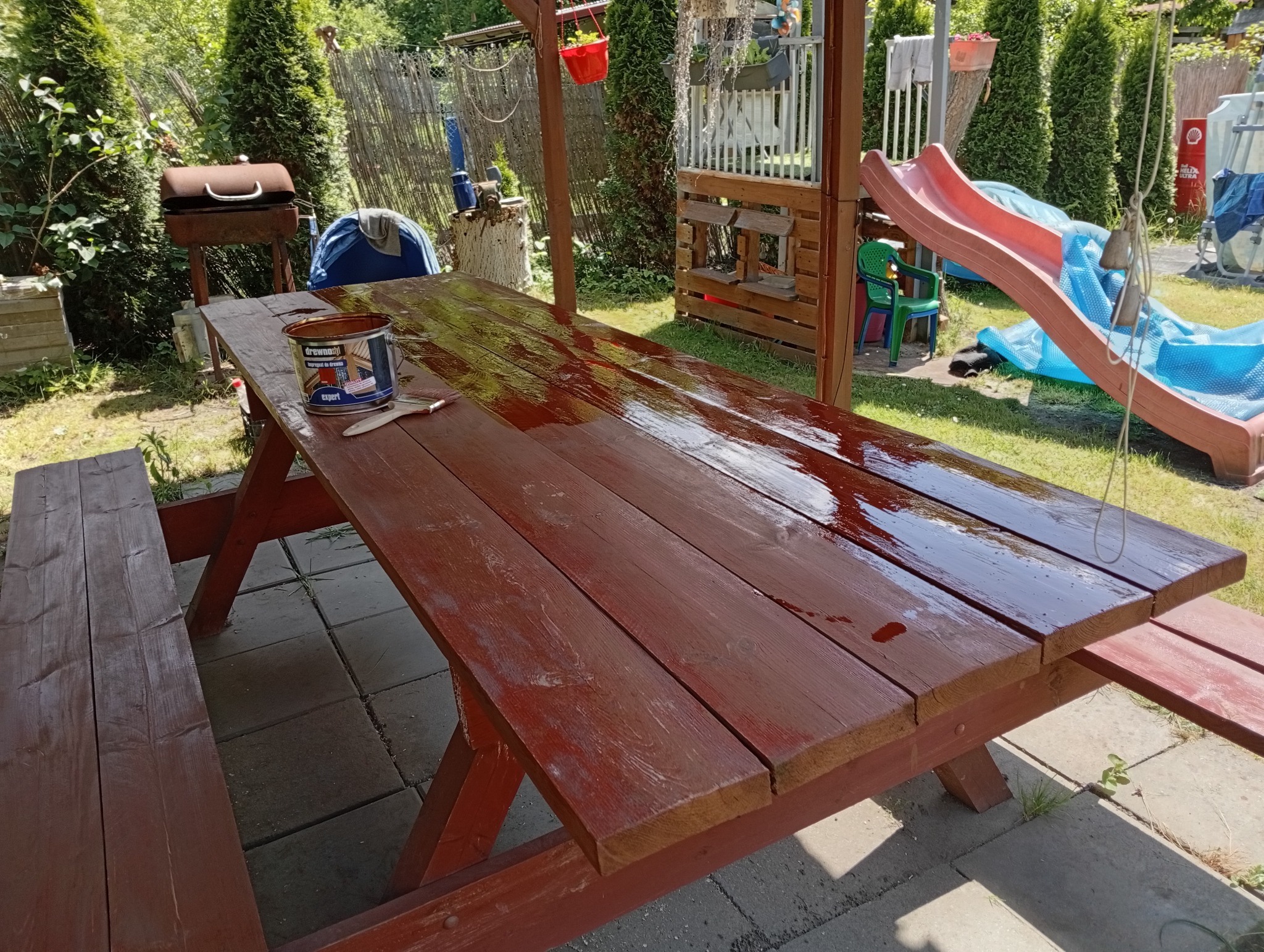 Impregnowany drewniany stół piknikowy w ogrodzie, częściowo pomalowany na brązowo. Na blacie puszka z impregnatem i pędzel. W tle zieleń i elementy placu zabaw.
