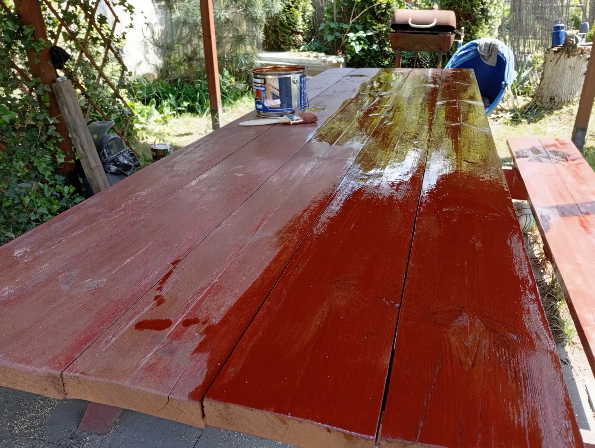 Malowanie drewnianego stołu piknikowego na zewnątrz. Część stołu pomalowana na czerwono, obok puszka farby i pędzel. Widoczne otoczenie: trawa, drzewa, grill.