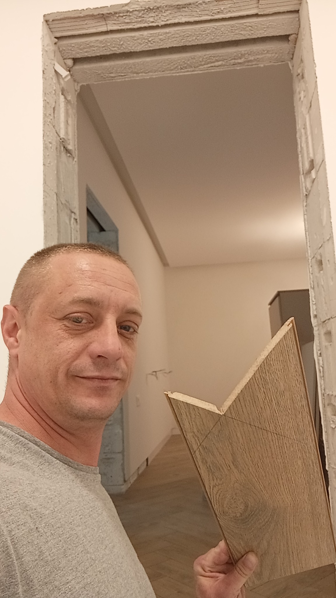 Mężczyzna trzyma panel podłogowy z narysowaną linią cięcia na tle odnowionego wnętrza z widoczną podłogą. Fragment futryny w trakcie prac wykończeniowych.